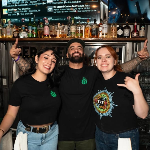Three smiling Servehzah Bottle Shop & Tap Room bartenders pose in front of a liquor shelf.