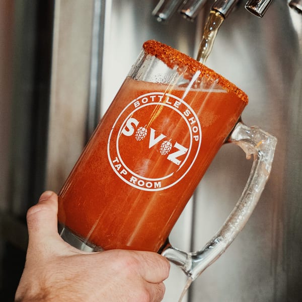 A hand holds a Servehzah Bottle Shop & Tap Room branded mug filled with a red drink.