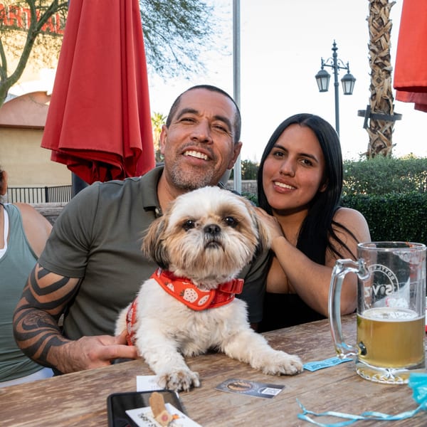 A man, woman, and small dog with a red bandana sit at an outdoor table with a beer.