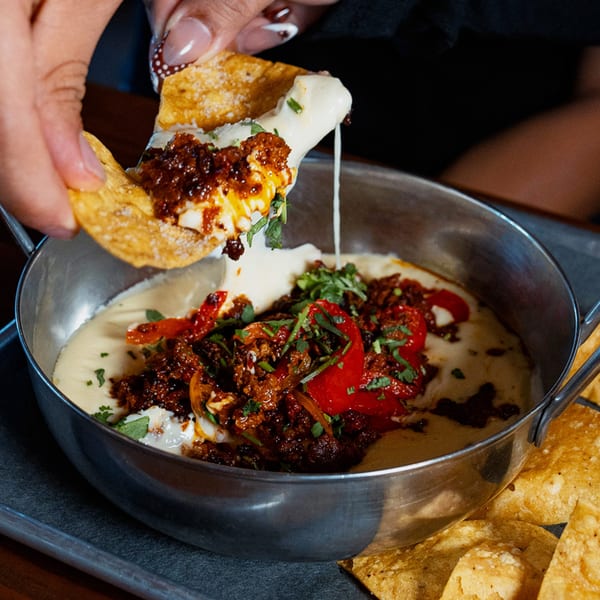 A hand dips a tortilla chip into a skillet of melted cheese, chorizo, and red peppers.