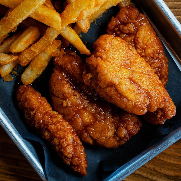 Crispy golden french fries and glazed chicken tenders served on black paper in a metal tray.