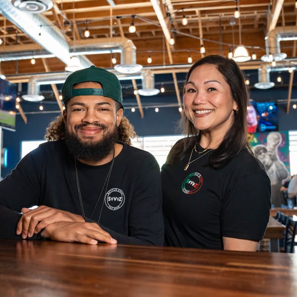 Two smiling Servehzah Bottle Shop & Tap Room staff members, a man and a woman, behind a wooden bar.