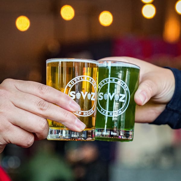 Two hands holding up Servehzah Bottle Shop & Tap Room branded glasses, one with golden beer, one with green.