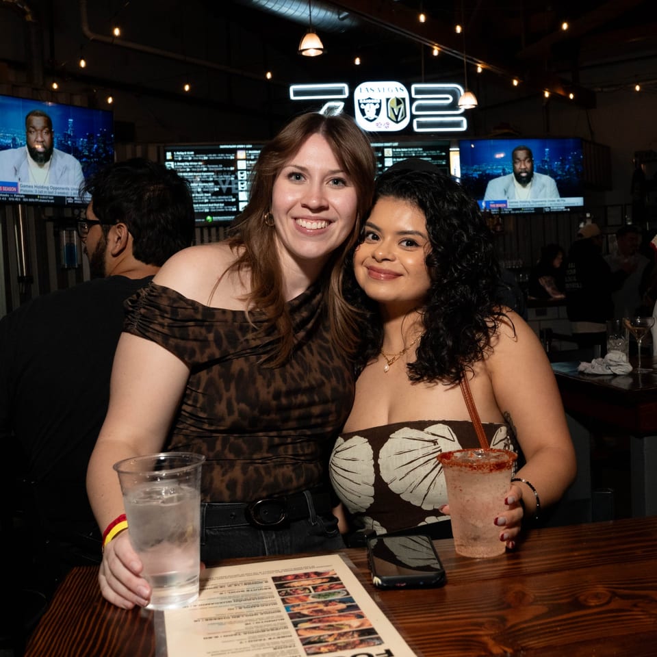 Two smiling women at a bar, one holding a drink, with TVs and a "702 Las Vegas" sign in the background.