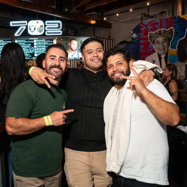 Three smiling men pose together at Servehzah Bottle Shop & Tap Room, with a neon "702" sign in the background.