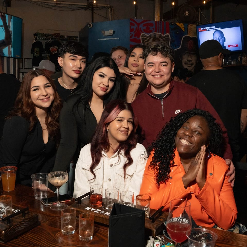 A group of diverse friends smiling and posing for a photo at a bar, with drinks on the table.
