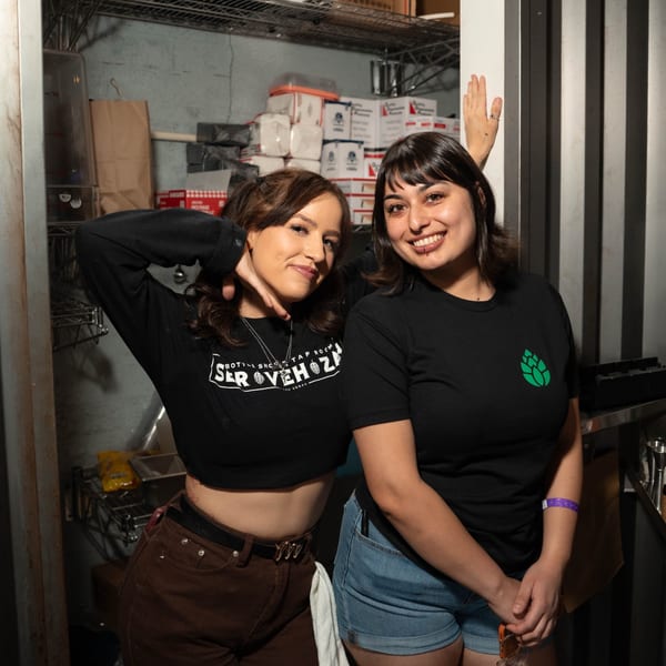 Two smiling Servehzah Bottle Shop & Tap Room employees pose in a storage room.