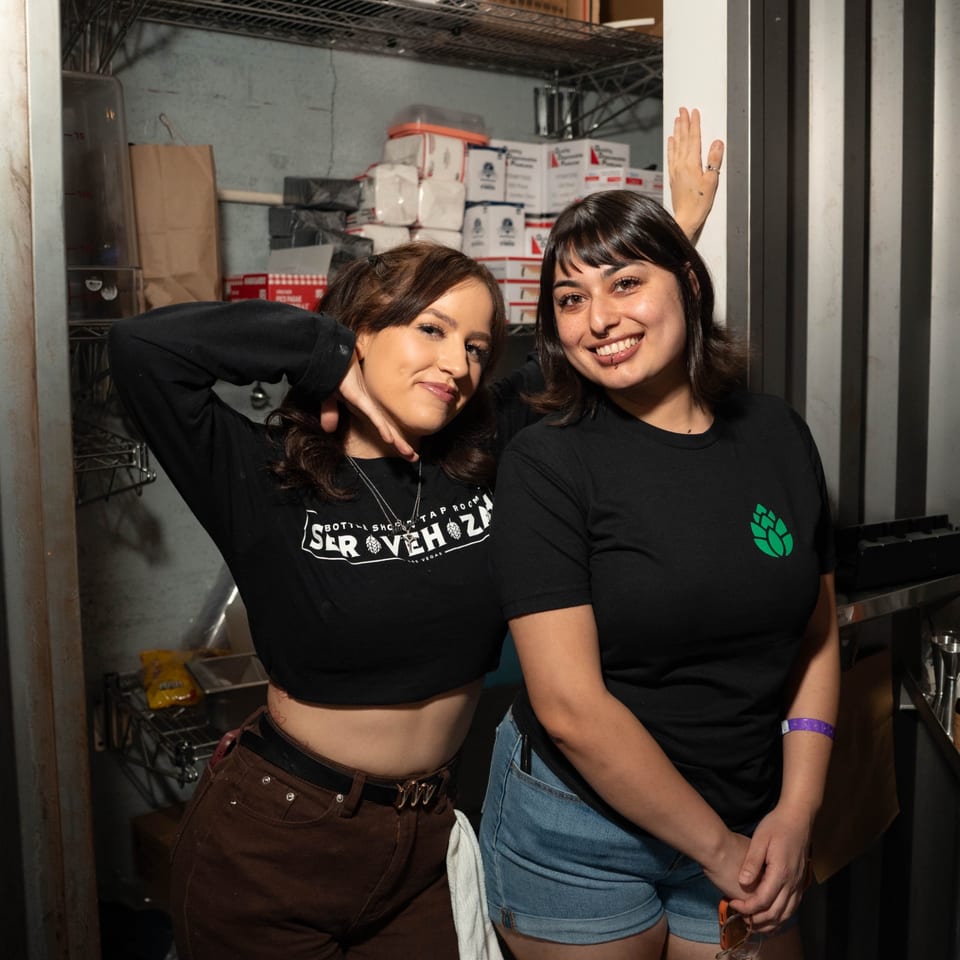 Two smiling Servehzah Bottle Shop & Tap Room employees pose in a storage area.
