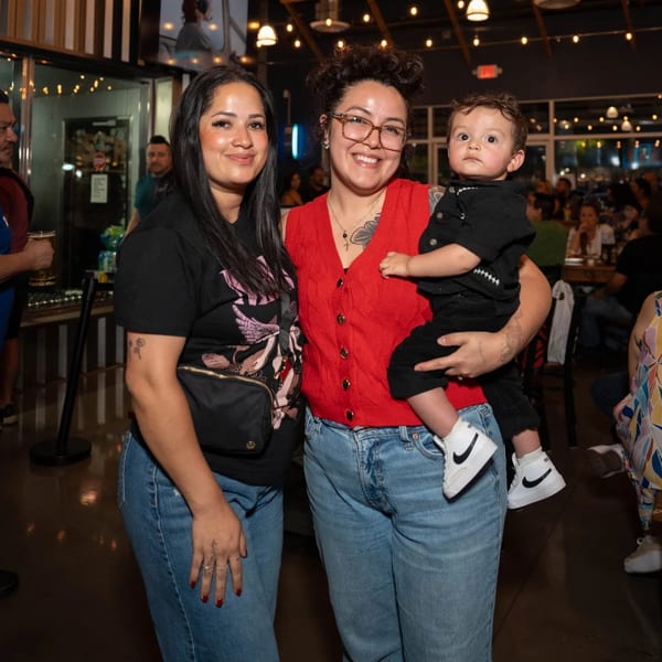Two women smiling, one holding a baby, inside Servehzah Bottle Shop & Tap Room.