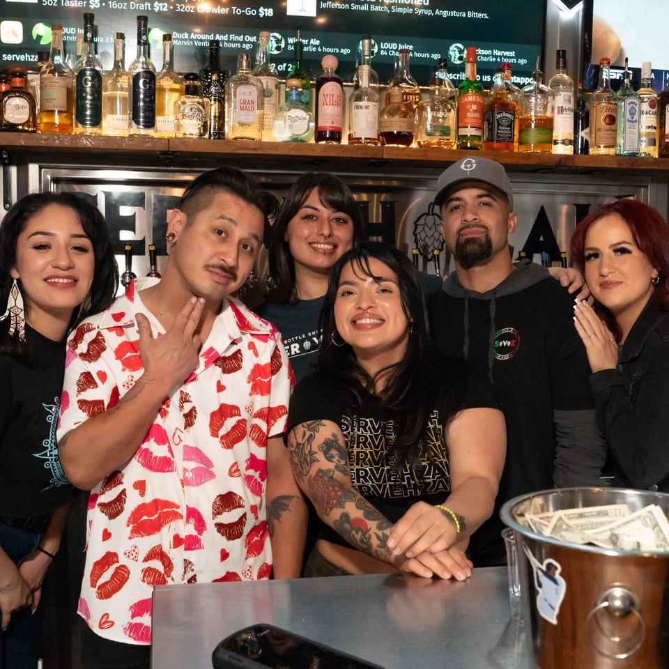 Six smiling Servehzah staff members pose behind a bar with bottles of liquor on shelves.