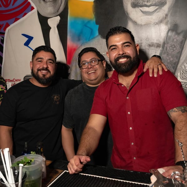 Three smiling men behind a bar, with a large mural of a man in a suit behind them.