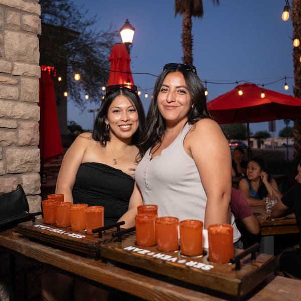Two smiling women at Servehzah Bottle Shop & Tap Room with a flight of orange drinks.