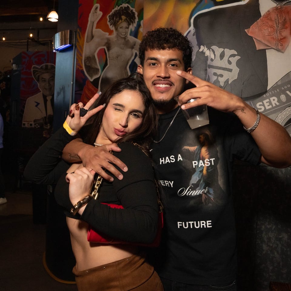 A smiling couple poses for a photo, making peace signs, in a dimly lit bar with murals.