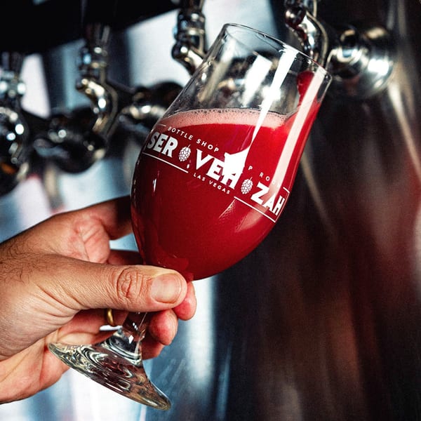 A hand holds a Servehzah branded glass as red beer pours from a tap.