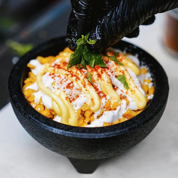 A hand in a black glove garnishes a bowl of elote with fresh cilantro at Servehzah.