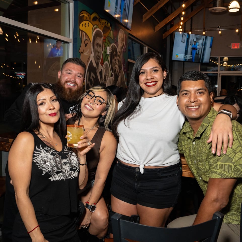 Five smiling friends pose together at Servehzah Bottle Shop & Tap Room, one holding a cocktail.