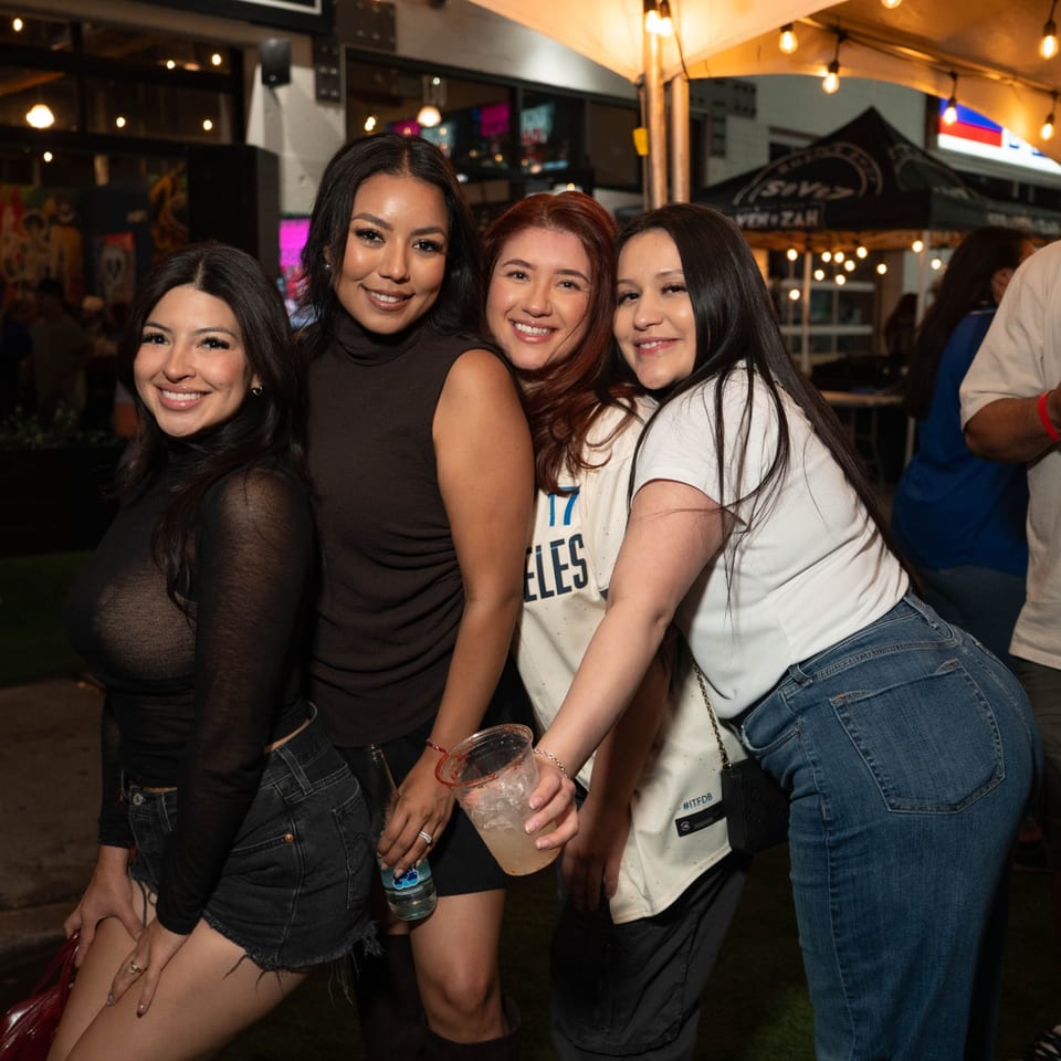 Four smiling women pose together at Servehzah Bottle Shop & Tap Room, holding drinks under string lights.