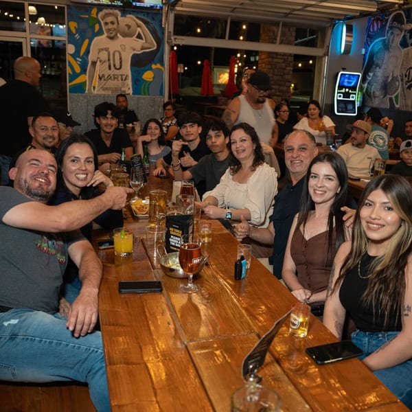 A large group of happy people sitting at a long wooden table in Servehzah Bottle Shop & Tap Room.