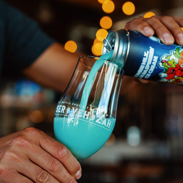 A person pours a vibrant blue drink from a can into a Servehzah branded glass.