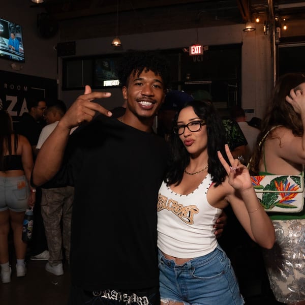 A man and woman pose for a photo, making peace signs at a lively bar.