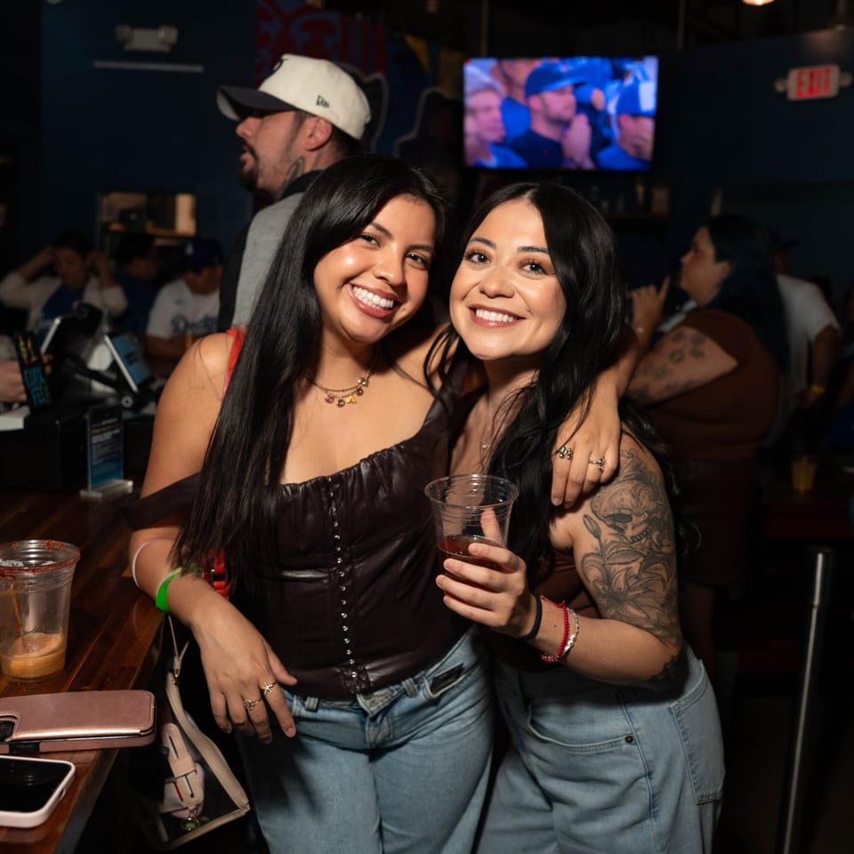 Two smiling women pose with drinks at Servehzah Bottle Shop & Tap Room, a bar.