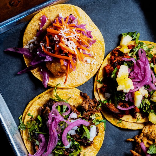 Three vibrant tacos with various fillings, including red cabbage, carrots, and pineapple, on a dark tray.