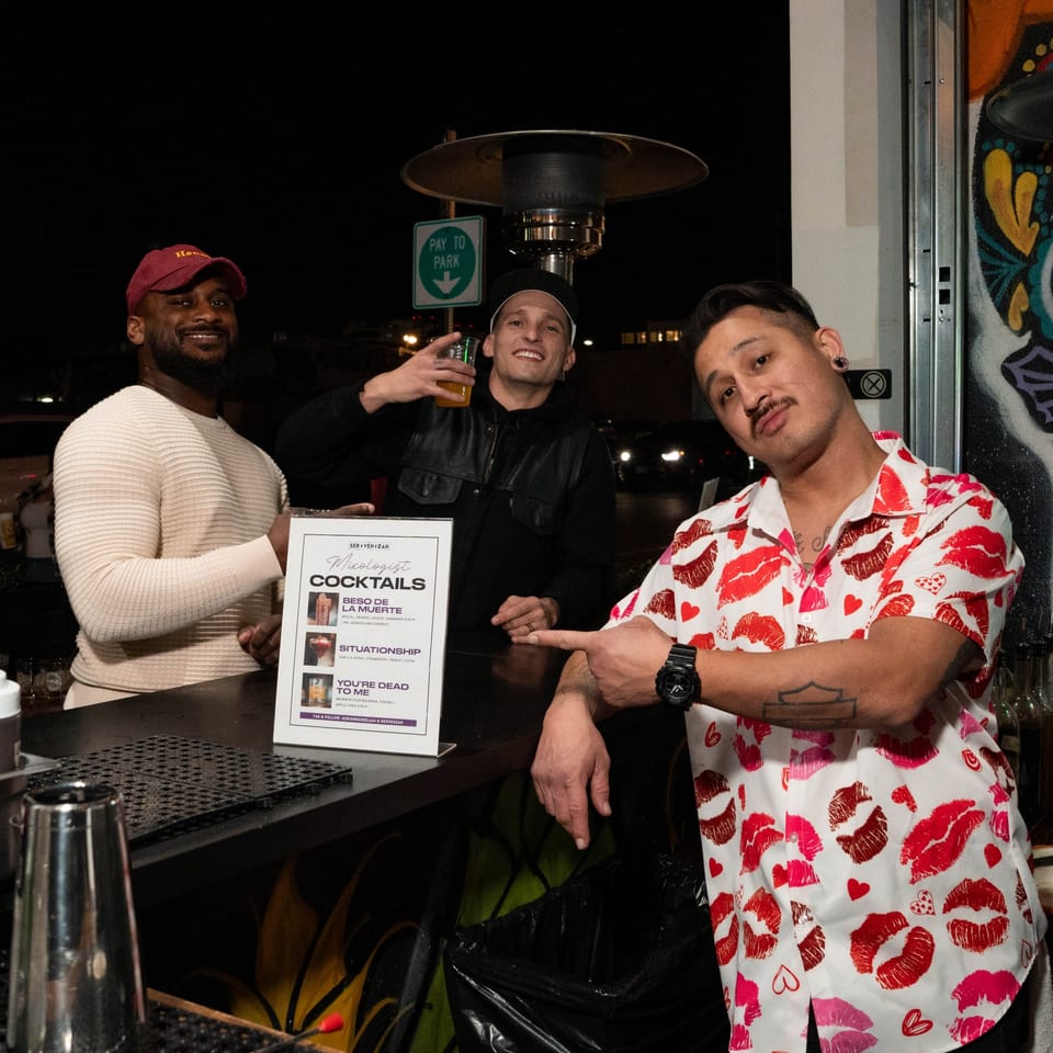 Three men smiling behind a bar with a cocktail menu, one pointing at the camera.