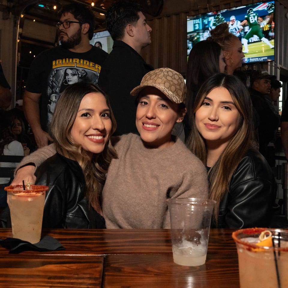 Three women smiling at a bar with drinks, a TV showing football in the background.