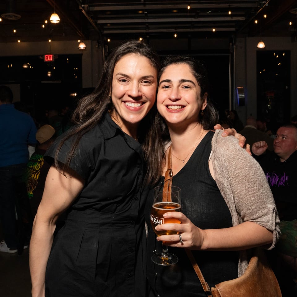 Two smiling women pose together, one holding a glass of beer at Servehzah Bottle Shop & Tap Room.
