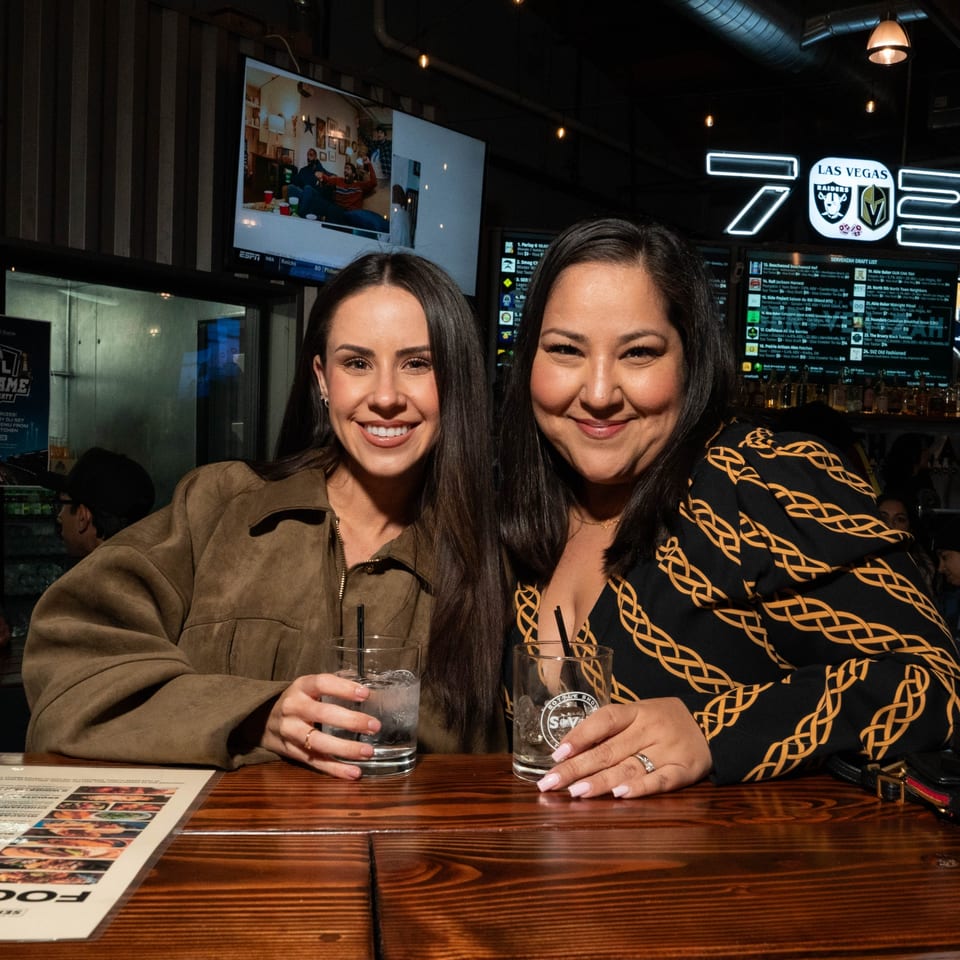 Two smiling women with drinks at a wooden bar counter in Servehzah Bottle Shop & Tap Room.