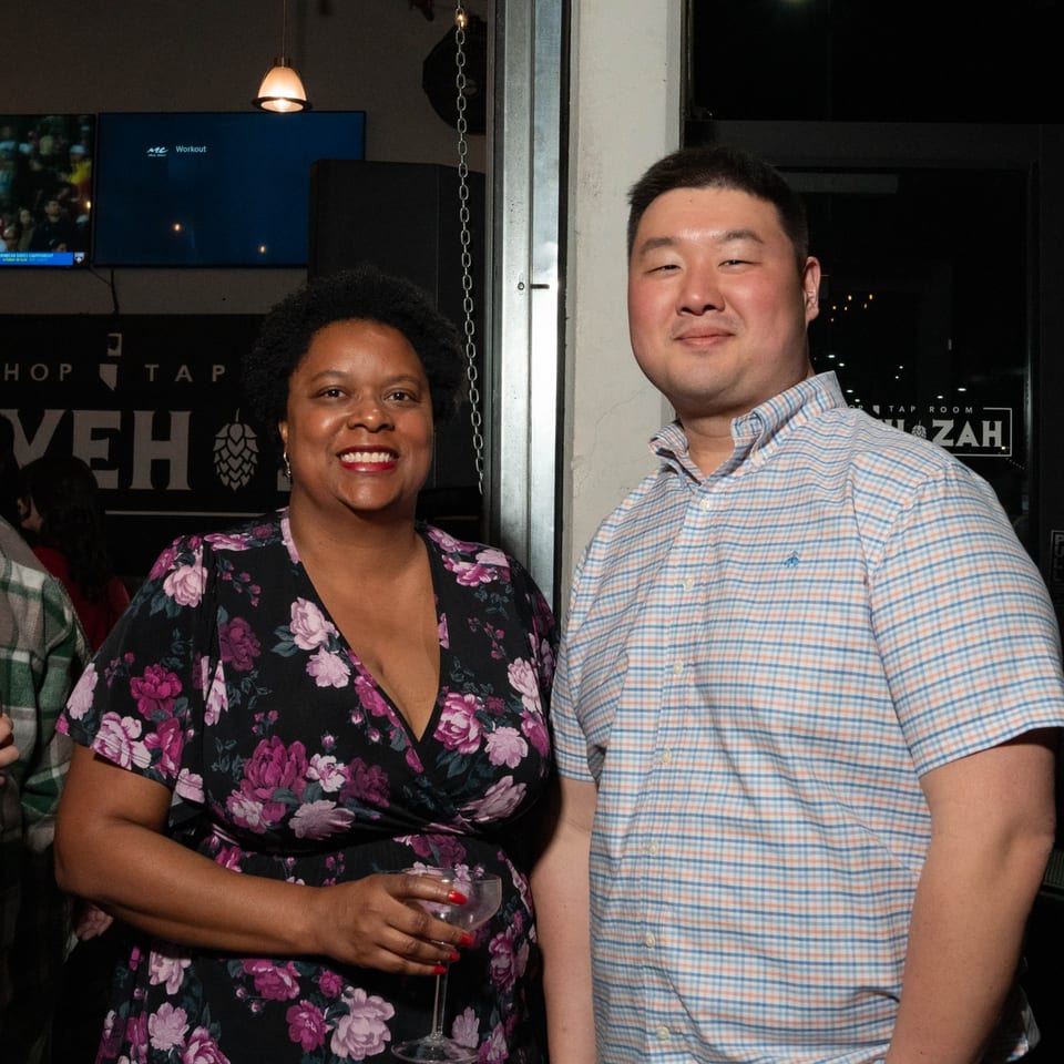 Two smiling people, a woman in a floral dress and a man in a plaid shirt, at Servehzah Bottle Shop.