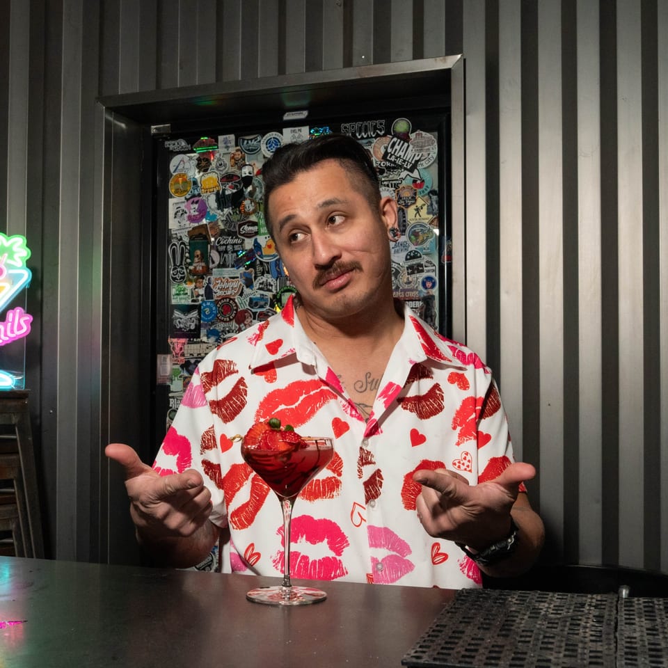 Bartender in a lip-print shirt shrugging behind a bar with a red cocktail.