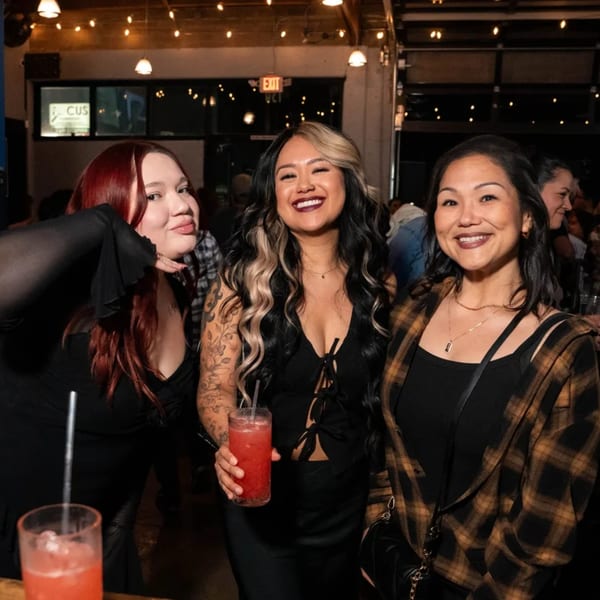 Three smiling women enjoying drinks at Servehzah Bottle Shop & Tap Room, with string lights in background.